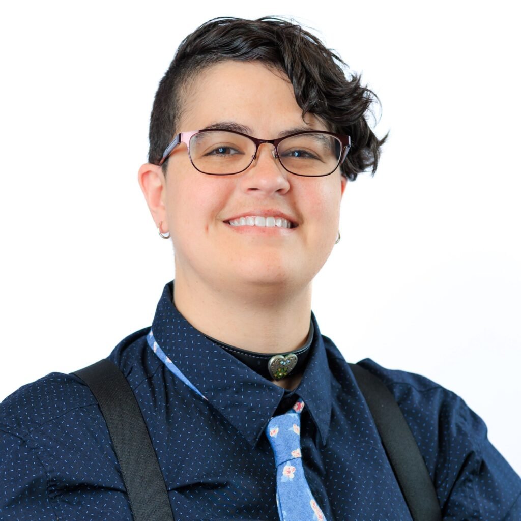 A nonbinary white person with glasses and short, dark hair. They are smiling at the camera while wearing a collared shirt and tie with roses.